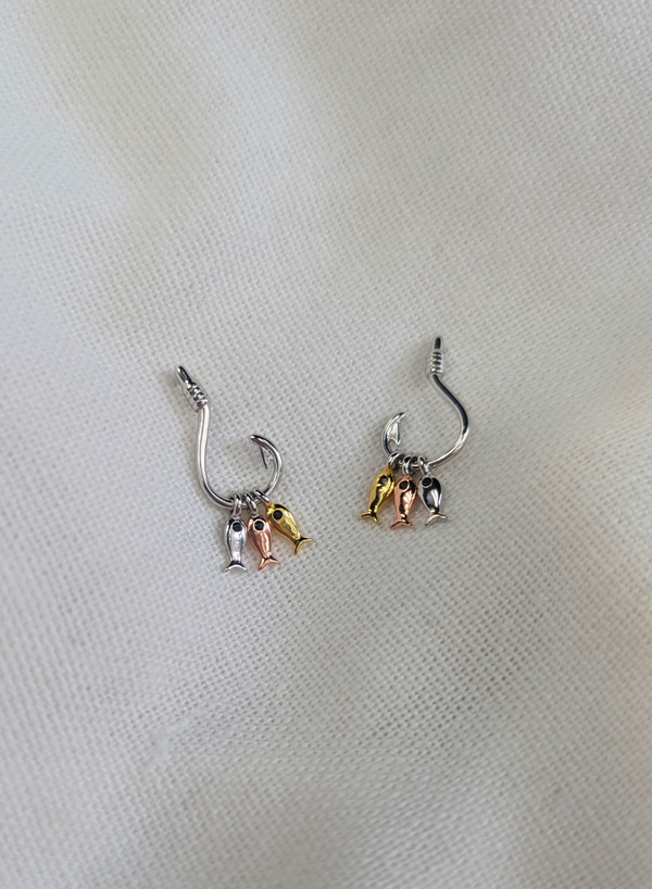 A pair of rhodium plated 925 sterling silver earrings with a unique hook design featuring three dangling fish, one silver, one bronze, and one gold. displayed on a white fabric surface.