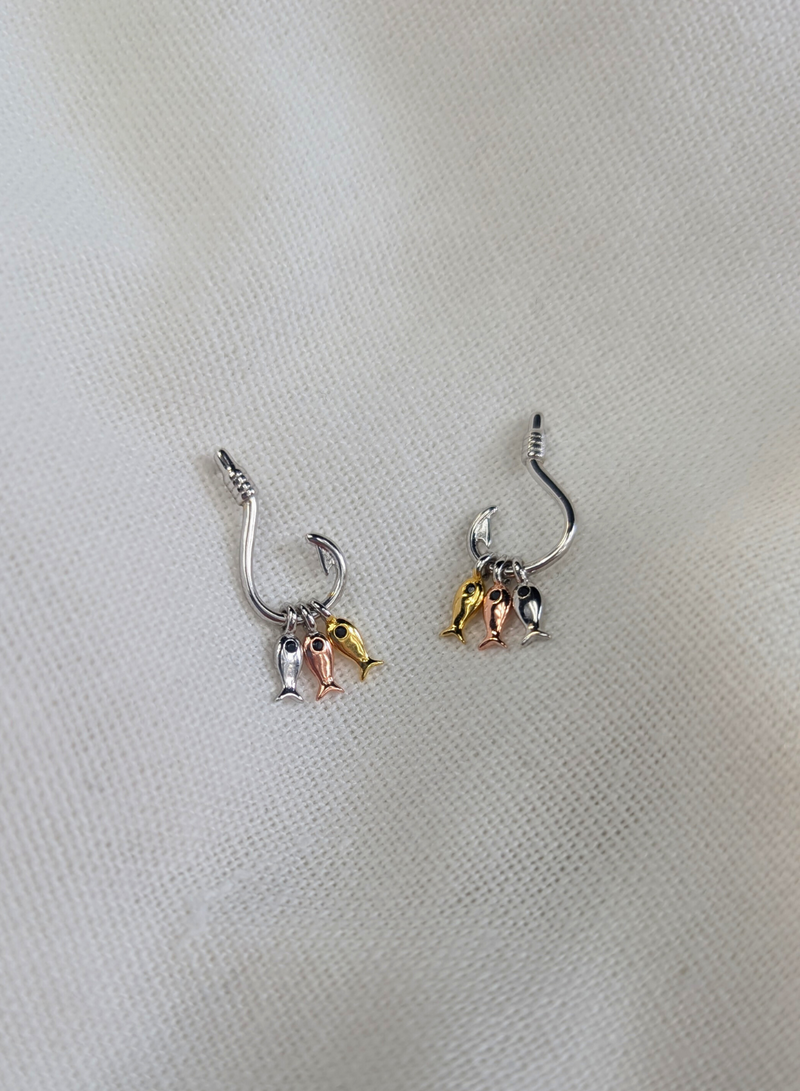 A pair of rhodium plated 925 sterling silver earrings with a unique hook design featuring three dangling fish, one silver, one bronze, and one gold. displayed on a white fabric surface.