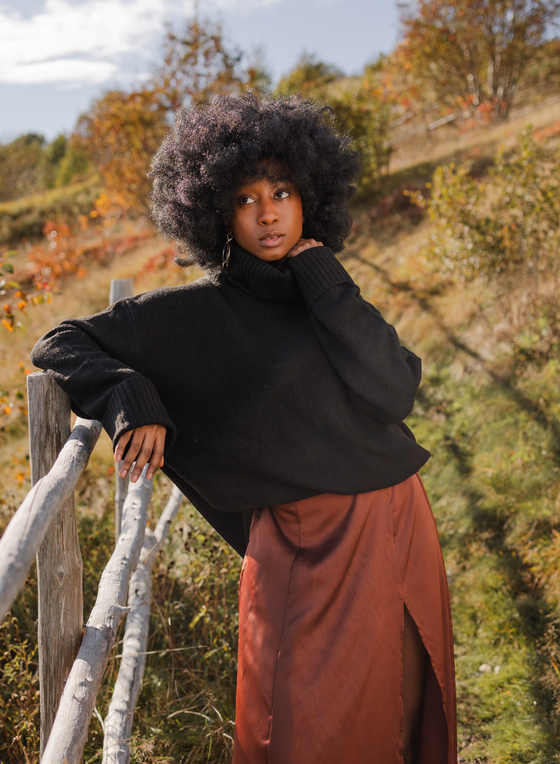 Model in Echo Verde Dog Walker Sweater and cinnamon slip skirt leaning against a wooden fence. 
