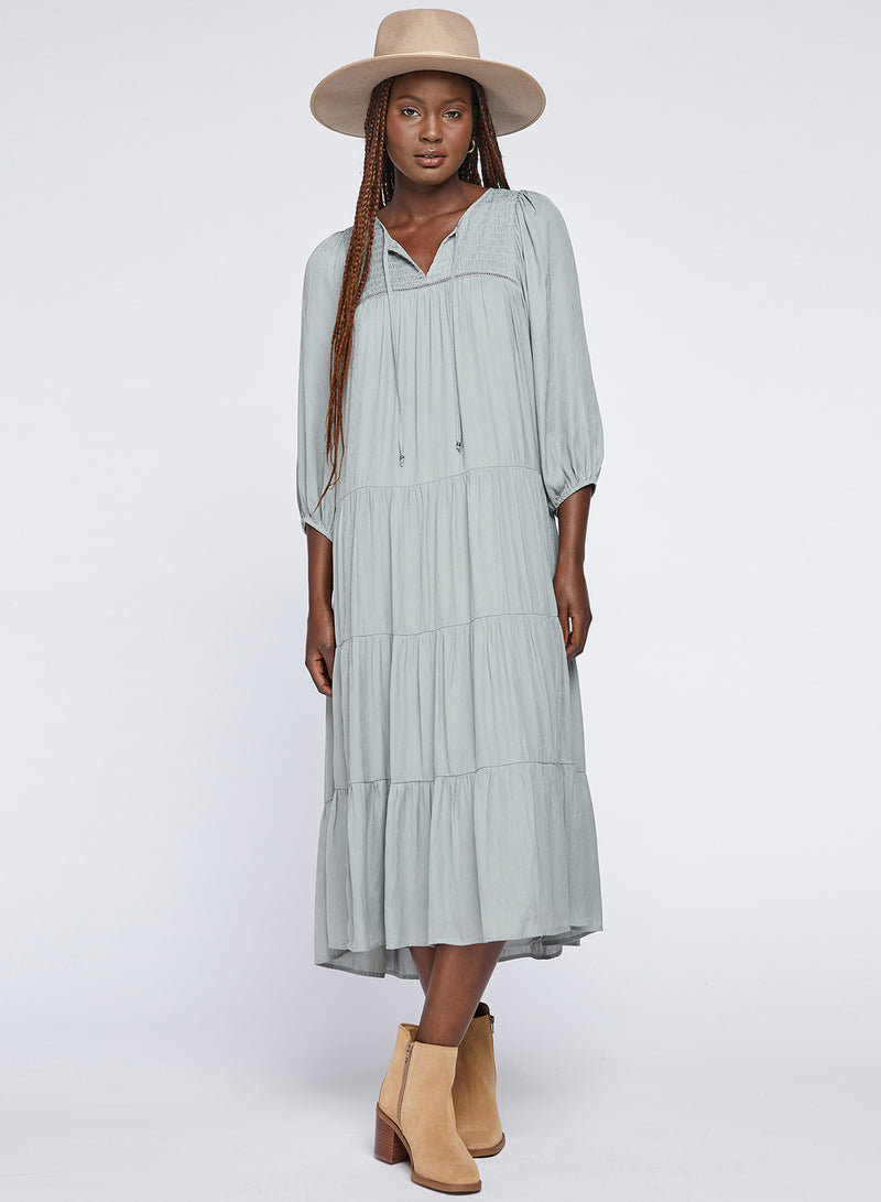 The photo shows a model wearing a midi length tiered dress, with slight gathering at each hemline. The garment also features loose three-quarter sleeves, a shirred panel at the bust, and drawstring closures at the neckline. The fabric is a soft, slightly shiny pale teal and is embossed with a faint feathered leaf motif