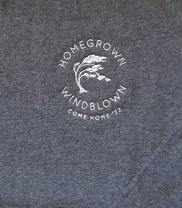 Close up of the embroidered "Homegrown Windblown" logo which is  surrounding an embroidered Tuckamore tree with the words Come Home '22 underneath, against a dark grey background, as it appears on the sweatshirt.